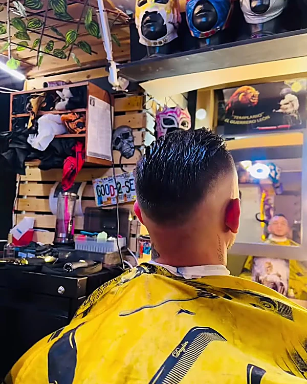 Nuca de un cliente con degradado bajo y cabello peinado hacia atrás, con capa amarilla estampada, frente a repisas con máscaras