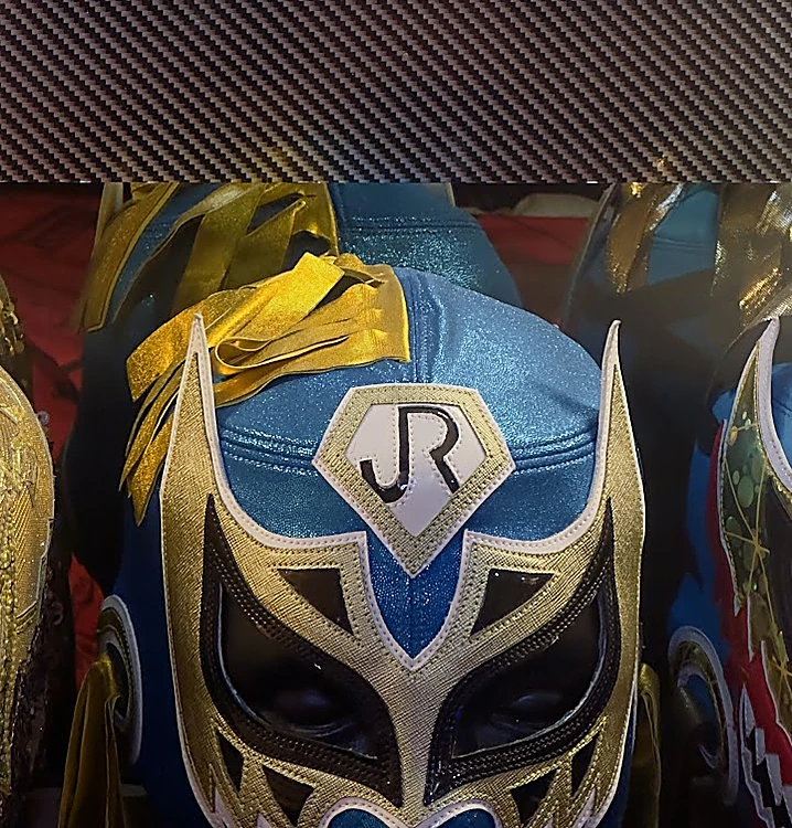 Máscaras de lucha libre de colores sobre el mostrador; al centro, una azul con plata y las letras JR en la frente