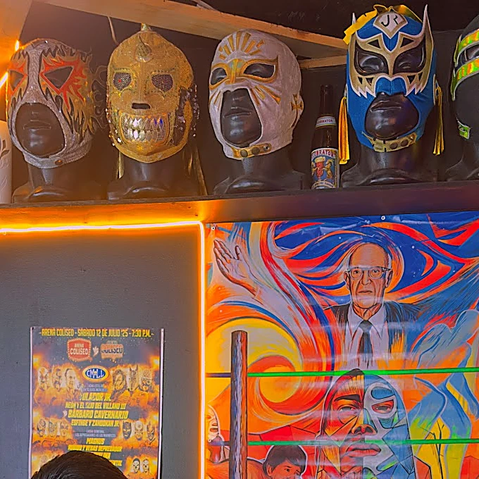 Fila de máscaras de lucha libre sobre una repisa con neón naranja y, debajo, el mural de colores del 80 aniversario del CMLL
