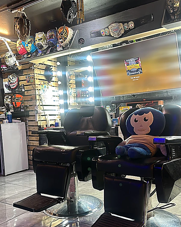 Dos sillones de barbero negros; en uno descansa un peluche de luchador azul. Arriba, máscaras en repisas de madera y un cinturón de campeonato